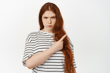 Upset young redhead girl complains, pointing right and frowning, look from under forehead with...