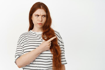 Angry teen redhead girl frowning, furrow eyebrows and pointing right at smth upsetting or strange, frustrated woman showing smth disappointing, standing against white background