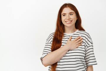 Obraz premium Image of happy girl with ginger hair, hold hand on heart and smiling, looking away with pleased and grateful face expression, thanking for smth, standing against white background