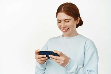 Young teen redhead girl playing video games on her phone, watching video live stream on cellphone, laughing and smiling, looking at mobile screen, white background