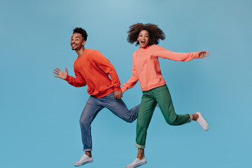 Fototapeta na wymiar Happy guy and girl in stylish outfits moving on blue background. Dark-skinned positive woman in green denim pants and man in jeans run on isolated