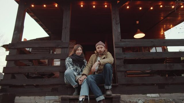 Portrait of young Caucasian couple in outerwear sitting on stairs of wooden terrace decorated with garland, holding hands and posing for camera