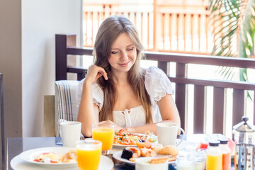 Obraz na płótnie Canvas Young Smiling Woman Sitting on Breakfast with Plates Full of Food - Fresh Omelette, Vegetables, Fruits and Orange Juice in Glass. Morning Food in Luxury Hotel