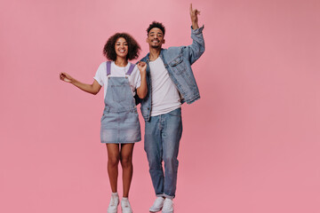 Obraz premium Brunette man and his girlfriend in jeans happily posing on pink background. Cool guy in denim jacket and girl in jumpsuit dance and smile
