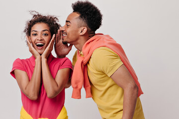 Dark-skinned man tells his girlfriend secret and she looks surprised into camera. Brunette guy in yellow tee and woman in red shirt gossip on isolated