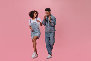 Full length portrait of couple in denim outfits dancing on pink background. Joyful man in jeans and brunette lady in blue dress move and jump on isolated