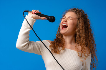 Young joyful curly-haired woman singing in microphone against blue background