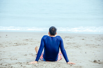 Young man sitting on the sand in a bathing suit, facing the wave, meditating, thinking