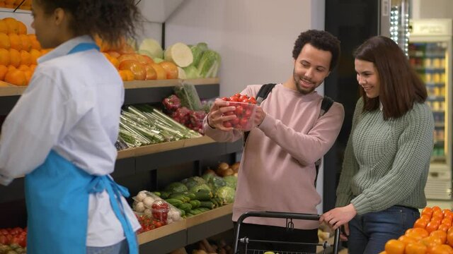 Mixed race woman supermarket employee working at store stand with shelves full of vegetables and fruits. Positive multinational couple of shoppers standing next choosing box of fresh cherry tomatoes
