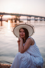 Fototapeta premium sunset picnic at the bridge over the river, Caucasian relaxed woman in a white dress and hat. 