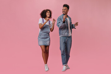 Cool girl and guy in jeans dance on isolated background. Attractive woman in denim dress and handsome man in white tee move on pink backdrop