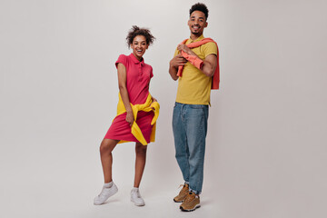 Fototapeta premium Full length portrait of man and woman in colorful outfits posing with smile on isolated background. Happy brunette girl in red dress and guy in jeans look into camera