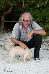 A handsome smiling long haired man with sunglasses is at beach petting small dog