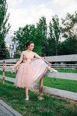 Fototapeta premium a young, elegant ballerina has lifted her foot on the wooden board of the fence of the horse pen in the amusement park and is adjusting the satin ribbons on her ballet flats.