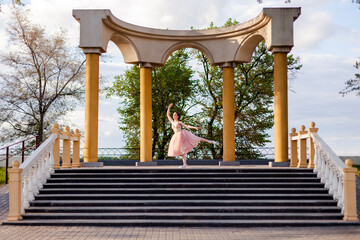 a ballerina dances with her leg raised in an arch of columns in urban architecture on the embankment of a river or sea, on a sunny day. 