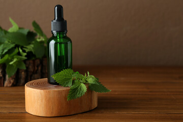 Glass bottle of nettle oil with dropper and leaves on wooden table against brown background, space for text