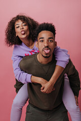 Obraz premium Brown-eyed man and woman having fun and showing tongues on pink background. Portrait of happy guy in brown outfit and girl hugging on isolated