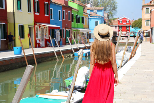 Back view of woman with red dress and hat walking along a channel on a sunny day in Burano Venice, Italy