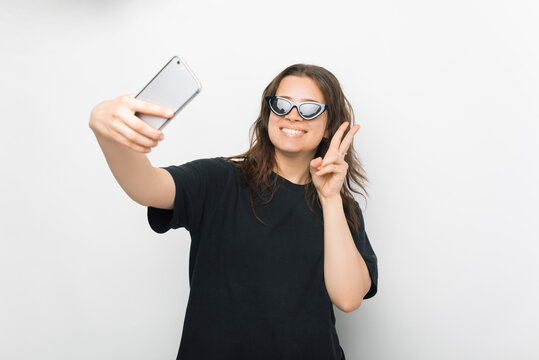 Portrait of a smiling young woman wearing sunglasses making selfie photo on smartphone, on a white background