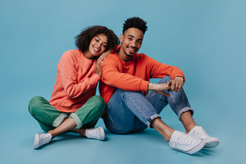 Fototapeta na wymiar Young man and woman smiling and sitting on blue background. Portrait og guy in red sweatshirt and woman in gree jeans chilling on isolated