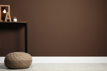 Comfortable knitted pouf and table with decor elements near brown wall indoors, space for text. Interior design