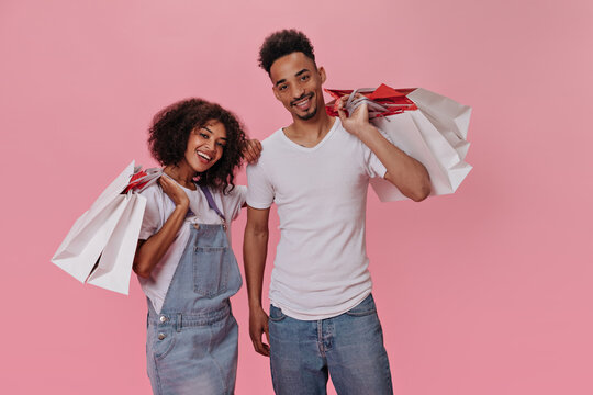 Guy and girl posing with shopping bags on pink background. Joyful curly woman in denim dress and happy man enjoy purchases