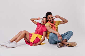 Fototapeta na wymiar Sitting on white background man and woman showing peace signs. Pretty dark-skinned girl in red dress and guy in yellow tee smile on isolated