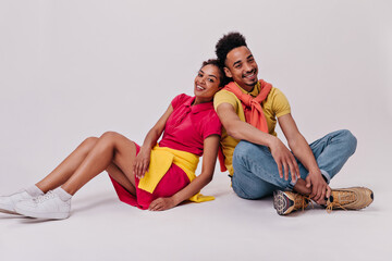Fototapeta na wymiar Man and woman in good mood sitting on white background. Cheerful girl in red dress and brunette guy in jeans smile on isolated