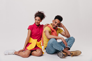 Charming lady and her boyfriend sit on white background. Pretty brunette girl in red dress and man in jeans pose on isolated backdrop