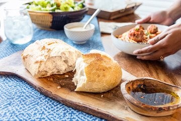 Rustic Italian bread torn in half in the foreground. Bread sits on a wooden cutting board with a bowl of olive oil and vinegar next to it.  Hands place a bowl of spaghetti and meatballs on the table.