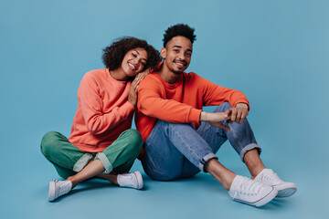 Charming couple in love sitting on blue background and hugging. Joyful woman in orange sweater and man in jeans widely smile on isolated