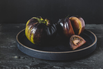 Two heirloom tomatoes on a black plate sitting on a slate surface with dramatic, moody lighting. The one on the right has a slice of tomato cut out from it. 