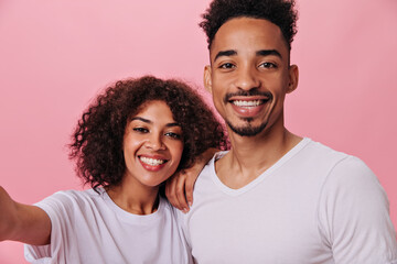 Positive Girl and guy in white T-shirts taking selfie. Dark-skinned happy woman and brunette man in tee smile and pose on pink background