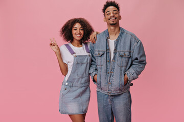 Couple in denim outfits posing on pink background. Dark-skinned woman and brunette man in white tees smiling on isolated