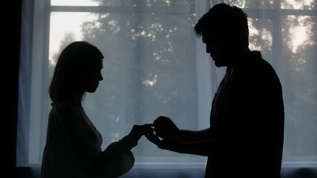 Silhouettes of man and woman, groom putting engagement ring to the bride in the evening at home, making proposal, young happy couple. Romantic view.