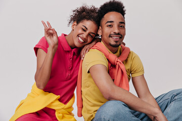 Naklejka premium Woman in red dress shows peace sign, winks and hugs her boyfriend. Happy lady in shirt and young man in jeans smile on white background