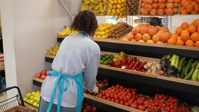 Back view of curly mixed race female grocery store employee in medical protective mask and gloves placing tomatoes. Woman tanding at rack with shelves full of fresh organic vegetables and fruits