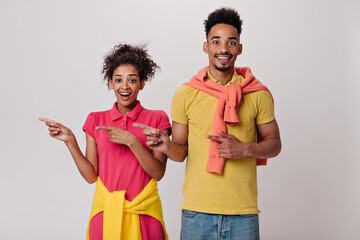 Surprised man and woman in colorful T-shirts point to place for text on isolated background. Dark-haired girl in red dress and guy in yellow tee smile on white backdrop