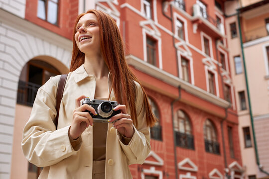 Caucasian Redhead Female Tourist In Beige Coat Standing On Street Of Old City And Shooting On Camera, Walking In Historical Places Alone, Enjoying Solo Trip. Portrait Of Cute Lady With Camera In Hands