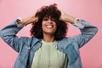 Positive woman in denim jacket and green t-shirt ruffles her hair against pink background. Happy...