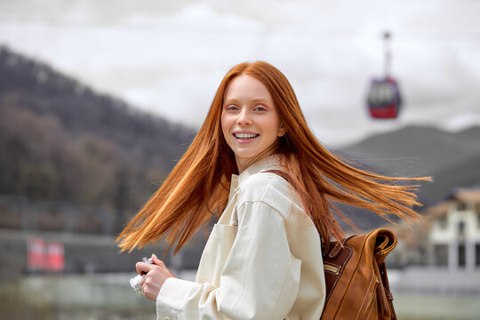 Woman with long red straight hair wearing beige coat jacket standing among mountains landscape, look at camera, turning head back. Copy space. Portrait