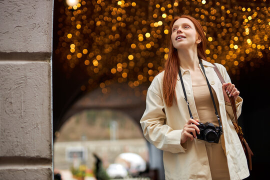 Pretty trendy young redhead woman with camera walk in city, boce garlands in the background. Portrait of beautiful lady in beige jacket enjoy walking alone outdoors. People lifestyle concept