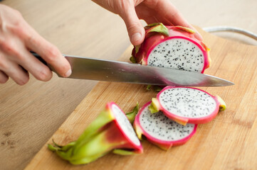Female hands is cutting a dragon fruit or pitaya with pink skin and white pulp with black seeds on wooden cut board on the table. Exotic fruits, healthy eating concept