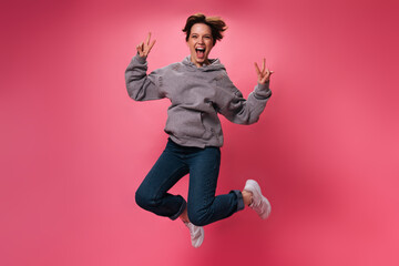 Pretty woman jumping and showing peace signs on pink background. Cheerful girl in grey hoodie and jeans dancing and smiling on isolated