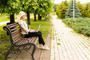 Fototapeta premium Young attractive businesswoman using a pc laptop computer while sitting on a wooden bench in a city park.
