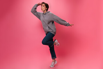 Woman in street style outfit looks into distance on pink background. Active teen girl in jeans and hoodie jumping on isolated