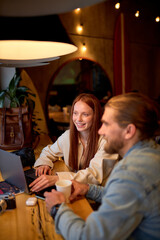Side view portrait of cheerful caucasian managers photographers redhead female and bearded guy working in cafe, use laptop, surfing internet, having talk, copy space. freelance concept