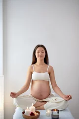 Schilderijen op glas Pregnant asian woman relax doing yoga, sitting in lotus position in spa cabinet alone with candles, with eyes closed, keep calm. Health lifestyle pregnancy beauty, interior. Mom-to-be with naked tummy © alfa27