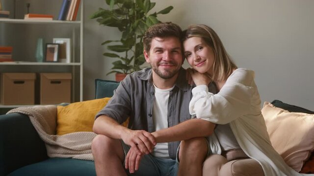 Happy family portrait. Young couple, lovely husband and wife sitting on sofa in living room. Cheerful man and woman smiling and posing on couch at home, looking in camera together and hugging. 