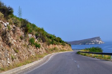 Winding road along Mediterranean coastline against blurred background of rock island among blue sea and blue sky.